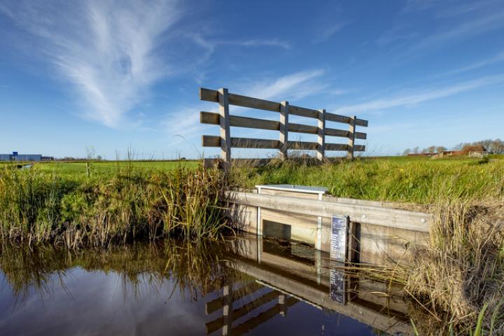 Foto van landschap met op de voorgrond een hek en een sloot met waterpeilaanduiding tegen de beschoeiing